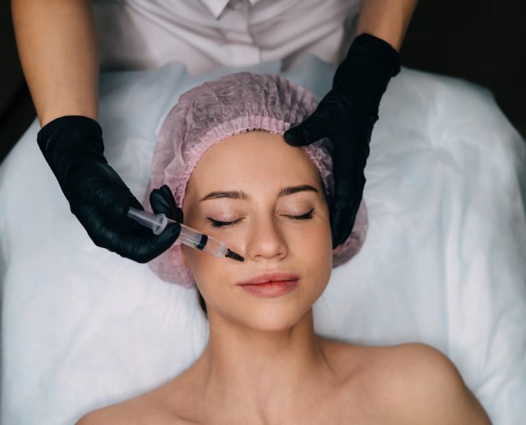 Cropped image of beautician doing botox injection with syringe to girl in beauty salon. Top view of caucasian woman client with closed eyes wear medical cap. Beauty concept. Female master wear gloves